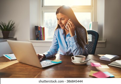Woman working on laptop at office while talking on phone, backlit warm light