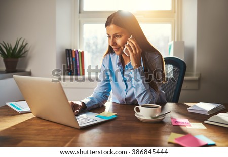 Photo of Woman working on laptop at office while talking on phone, backlit warm light
