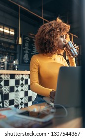 Woman Working On Laptop Sitting At A Restaurant Drinking Coffee. Afro American Woman Enjoying A Cup Of Chocolate Shake While Working On Laptop At A Cafe.