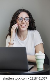 Woman Is Working On A Laptop, Talking On The Phone And Smiling. A Happy Successful Spanish Woman Entrepreneur Or Student Works In A Freelance Coworking. Marketer And Social Media Manager