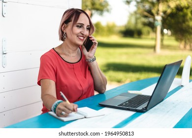 Woman working on modern laptop outdoor against a white wooden background - 40 years old woman calling while taking notes - Freelancer or female entrepreneur working with her devices from remote