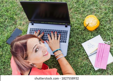 Woman working outdoors with laptop - Smiling female entrepreneur sitting on lawn typing on pc during coronavirus lockdown - Health orange juice for break. Remote work concept into the nature. Top view