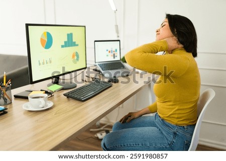 Woman working remotely experiencing sharp neck pain, massaging cervical spine while sitting at home workspace with poor ergonomic setup