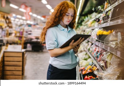 Woman Working In A Supermarket. Female Assistant Using A Digital Tablet For Taking Inventory Of Products In Display Rack In A Grocery Store.