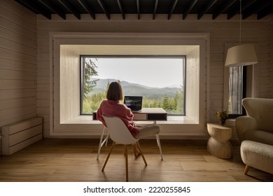 Woman works on laptop while sitting by the table in front of panoramic window with great view on mountains. Wide interior view. Remote work and escaping to nature concept - Powered by Shutterstock