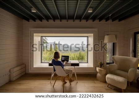 Woman works on laptop while sitting by the table in front of panoramic window with great view on mountains. Wide interior view. Remote work and escaping to nature concept