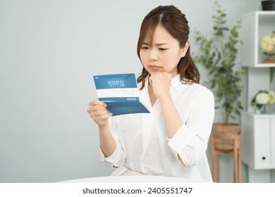 A woman worries about budget management with a savings passbook in hand.The book in the woman's hand is a bank book with the name of a fictitious Japanese bank.