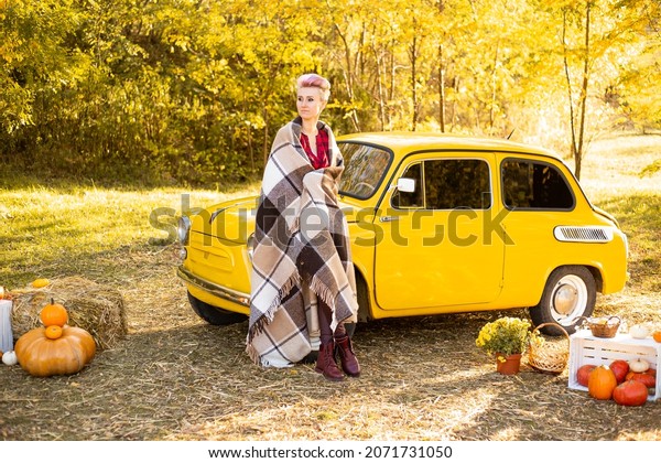 woman wrapped in a blanket\
near yellow retro car in autumn flowers background with golden\
trees