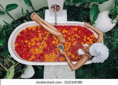 Woman with wrapped towel on head is drinking tea or coffee while relaxing in outdoor bath with flowers in Bali spa hotel, top view from above. - Powered by Shutterstock