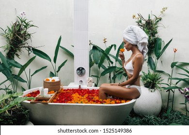 Woman With Wrapped Towel On Head Is Relaxing In Outdoor Bath With Flowers In Bali Spa Hotel.