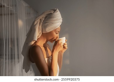 A woman wrapped in a towel with a towel on her head enjoys a cup of coffee in the morning light. The serene setting captures a moment of relaxation and self-care, perfect for a peaceful start to the