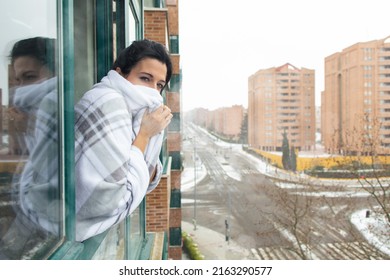 Woman Wrapped In A Warm White Blanket Looking Out The Window During A Cold Winter Snow Storm.