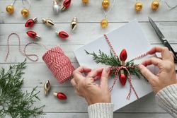 Woman Wrapping Christmas Gifts With Red And White Bakers Twine, Greenery And Red Light Bulbs, Flat Lay