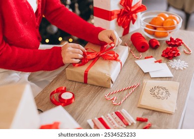 A woman wrapping Christmas gifts with red ribbon, surrounded by festive decorations, candy canes, and oranges on a table. Capturing the essence of holiday preparation and seasonal joy. - Powered by Shutterstock