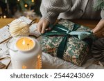 Woman wrapping stylish christmas gift on rustic table with modern paper ornaments and lantern on background of christmas tree lights bokeh. Merry Christmas! Atmospheric christmas time