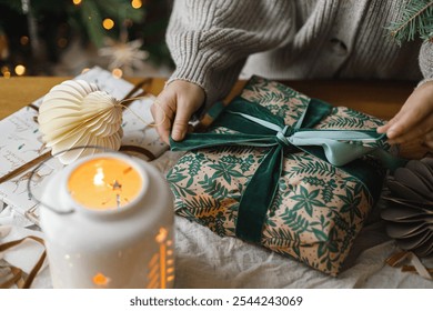 Woman wrapping stylish christmas gift on rustic table with modern paper ornaments and lantern on background of christmas tree lights bokeh. Merry Christmas! Atmospheric christmas time - Powered by Shutterstock