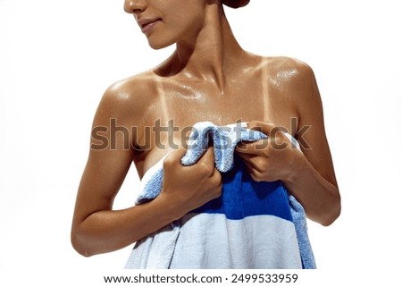 Woman wraps herself in towel, revealing stark tan lines on her deeply tanned skin against white studio background. Concept of beauty, body care, dermatology, summer season, protection.