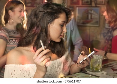 Woman Writes A Message On The Mobile Phone And Smoking A Cigarette At The Counter Of A Club While Her Friends Are Talking In The Background