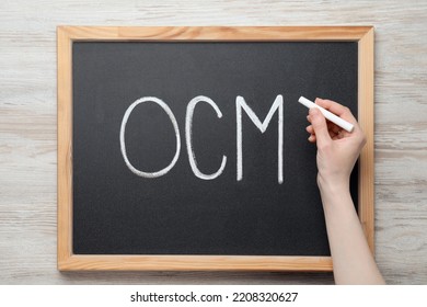 Woman Writing Abbreviation OCM (Organizational Change Management) On Small Blackboard With At White Wooden Table, Top View