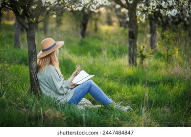 Woman is writing diary and resting in blossoming orchard. Mindfulness and reconnect the moment in spring nature