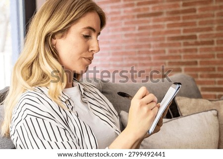 Woman writing notes in a notebook while sitting on a couch at home, planning and working casually with a pen, relaxed and focused in a cozy living room.