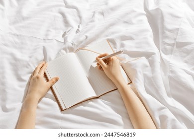 Woman writing on blank white page diary journal on white bedcloth in bed, holds paper notebook and pen in hands, device free time before sleep or morning, digital detox, psychological wellbeing - Powered by Shutterstock