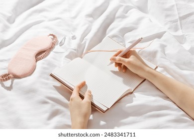 Woman writing on blank white page diary journal on white bedcloth in bed, holds paper notebook and pen in hands, device free time before sleep or morning, digital detox, psychological wellbeing - Powered by Shutterstock