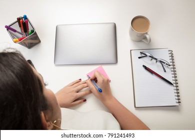 Woman Writing A Sticky Note Reminder For Herself As She Sits Working At A Desk With A Closed Laptop And Notepad With Pen And Glasses In An Overhead View
