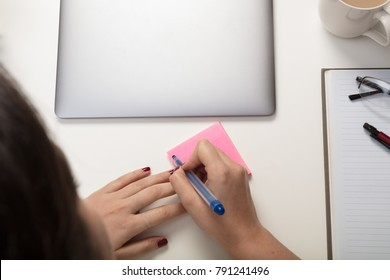 Woman Writing A Sticky Note Reminder For Herself As She Sits Working At A Desk With A Closed Laptop And Notepad With Pen And Glasses In An Overhead View