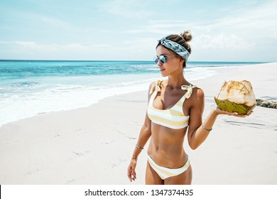 Woman in yellow bikini and sunglasses drinking fresh coconut juice while relaxing on sandy tropical beach. Healthy summer vegan diet concept.