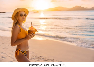 Woman in yellow bikini and sunglasses drinking fresh coconut juice while relaxing on sandy tropical beach. Healthy summer vegan diet concept. Copy space - Powered by Shutterstock