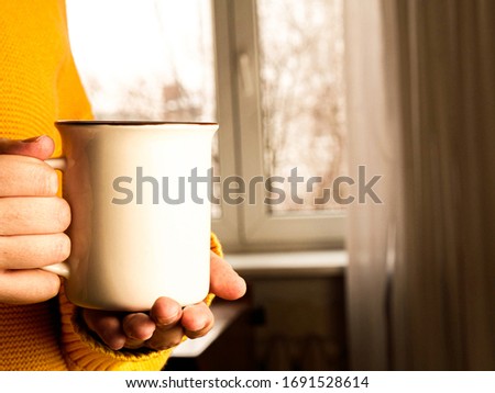 Woman in yellow cozy sweater holding red cup of hot drink on window background. Copyspace, close up, mockup, home atmosphere concept
