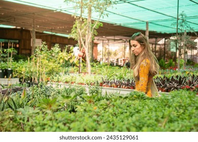 A woman in a yellow dress is delicately tending to an assortment of lush plants at a greenhouse during spring. The vibrant greenery around her suggests a verdant and thriving environment, likely full  - Powered by Shutterstock