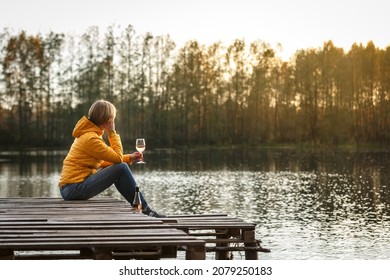 Woman in a yellow jacket relaxing on a wooden pier on a lake with a glass of rose wine watching fall sunset alone. Enjoying nature, relaxation and meditation