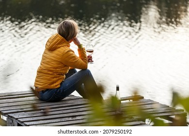 Woman in a yellow jacket relaxing on a wooden pier on a lake with a glass of rose wine watching fall sunset alone. Enjoying nature, relaxation and meditation