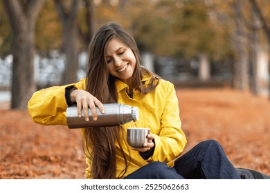 woman in yellow jacket sits comfortably in park among fallen autumn leaves. She smiles as she pours warm drink from stainless steel thermos into her cup. enjoying the fresh air. hot drink coffee tea  - Powered by Shutterstock