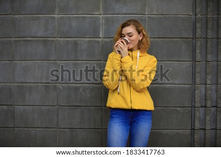 Woman in yellow jacket sniff aroma of takeaway coffee near grey wall