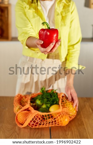 Woman in yellow jacket unpacking shopping mesh eco bag with healthy vegan vegetables and fruits on the kitchen at home, Healthy eating vegetarian concept.