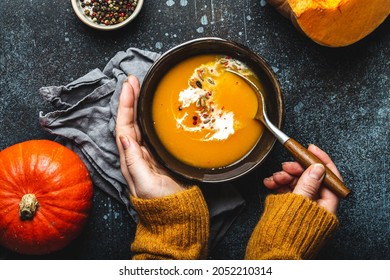 Woman In Yellow Knitted Sweater Eating A Pumpkin Cream Soup On Dark Stone Background With Spoon Decorated With Cut Fresh Pumpkin, Top View. Autumn Cozy Dinner Concept 
