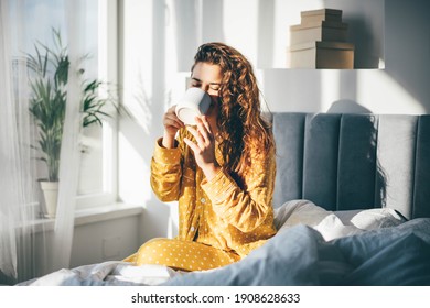 Woman in yellow pajama sitting on bed and drinking coffee at the sunny morning bedroom.  - Shutterstock ID 1908628633