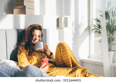 Woman in yellow pajama sitting on bed and drinking coffee and looking to the phone at the sunny morning bedroom. 
