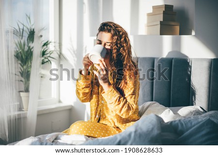 Woman in yellow pajama sitting on bed and drinking coffee at the sunny morning bedroom. 