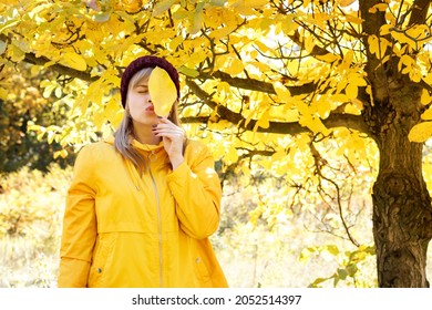 a woman in a yellow raincoat covers her face with an autumn leaf against the background of autumn leaves. free space for text