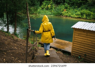 Woman in a yellow raincoat and rubber boots descends the stairs to the pier by the lake in a green wet forest during the rain. The concept of travel and tourism.
