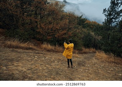 A woman in a yellow raincoat standing on a hill admiring scenic view while checking her cell phone - Powered by Shutterstock