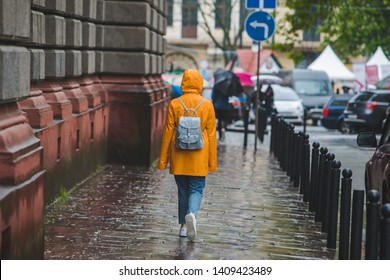 Woman In Yellow Raincoat Walking By City Streets Under Rain. View From Back