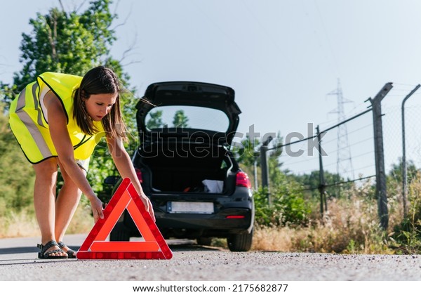 Woman with yellow reflective\
vest placing the emergency warning system for her broken down car.\
Young girl supporting a warning triangle for broken down\
vehicles.