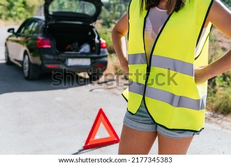 Woman in yellow reflective vest standing desperate with her broken car on the shoulder. Close up image of a young girl worried about her broken car while waiting for insurance assistance.