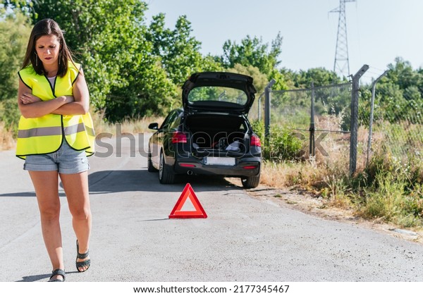 Woman with yellow reflective vest standing wandering
next to her broken car on the shoulder. Young girl walking by her
vehicle while waiting for insurance assistance with an emergency
triangle on.