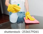 Woman in yellow rubber gloves holding a bottle of cleaning solution on a glass table with a rag